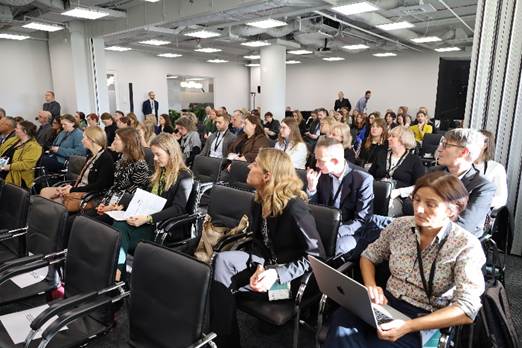 The image depicts a large, crowded conference room with attendees seated in rows of black chairs, some working on laptops, and a general atmosphere of professional engagement.

AI-generated content may be incorrect.