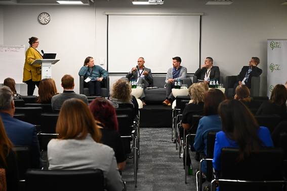 The image depicts a professional conference or panel discussion, with a diverse group of individuals seated in rows, some engaging in conversation, and a backdrop featuring a conference logo and various educational and organizational names.

AI-generated content may be incorrect.